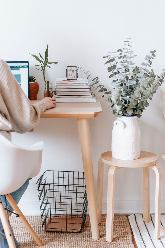 Crop unrecognizable female browsing netbook while sitting at wooden table with books in light room with green twigs at home